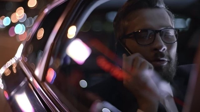 Close up of bearded businessman in glasses and suit talking on mobile phone while riding in backset of car at night
