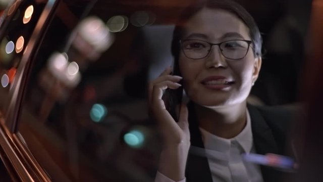Close up of Asian businesswoman in glasses and suit chatting on mobile phone and looking out window of moving car at night