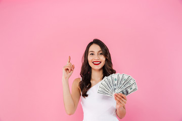 Gorgeous asian woman 20s holding fan of money in dollar cash and pointing finger upward on copyspace with smile, isolated over pink background