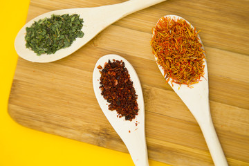 Plastic spoons with dry spices and fresh herbs on a wooden cutting board with yellow background, top view, close up