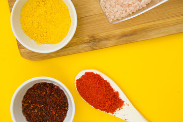 Plastic cups and spoons with dry spices and fresh herbs on a wooden cutting board with yellow background, top view, close up