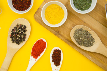 Plastic cups and wooden spoons with dry spices and fresh herbs on a wooden cutting board with yellow background, top view, close up