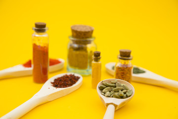 Glass bottles and spoons with dry spices and fresh herbs on a wooden cutting board with yellow background, top view, close up