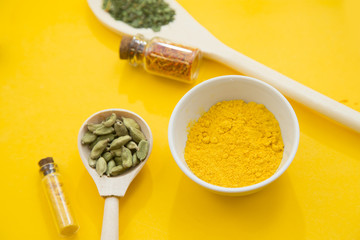 Glass bottles and spoons with dry spices and fresh herbs on a wooden cutting board with yellow background, top view, close up