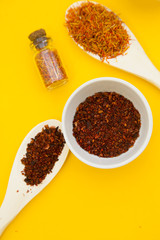 Glass bottles and spoons with dry spices and fresh herbs on a wooden cutting board with yellow background, top view, close up