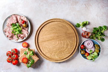 Ingredients for making sandwiches. Sliced beef meat, rye whole graine bread, green salad, tomatoes, pickled cucumber, onion on plates, empty wooden tray over grey texture background. Top view, space. © Natasha Breen