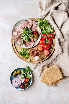 Ingredients for making sandwiches. Sliced beef meat, rye whole graine bread, green salad, tomatoes, pickled cucumber, onion on blue plates, wooden tray over grey texture background. Top view, space.