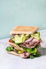 Beef and vegetables sandwiches with sliced meat, cucumber, green salad, rye whole grain bread in stack over grey green pin-up style background.