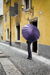 The figure of a girl in trousers closed umbrella color ultraviolet light against the yellow houses on the old Italian street. Above the girl's head there is an old yellow street lamp. Vertical photo.