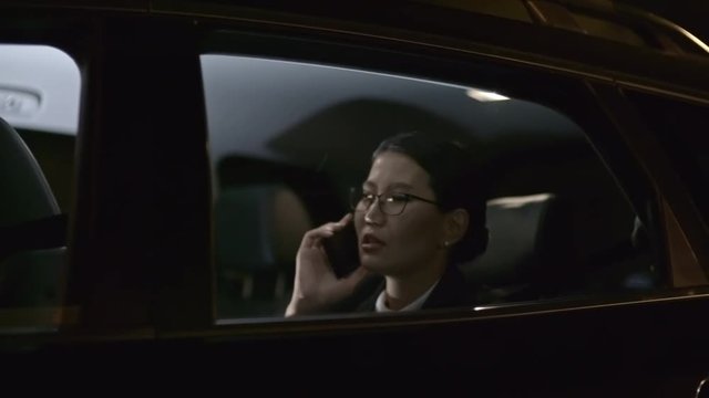 Tracking With Side View Of Asian Businesswoman In Glasses Talking On Mobile Phone While Riding In Backseat Of Moving Car At Night