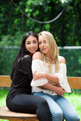 A pair of proud lesbian in outdoors sitting on a wooden table, brunette woman is hugging a blonde woman, in a garden background
