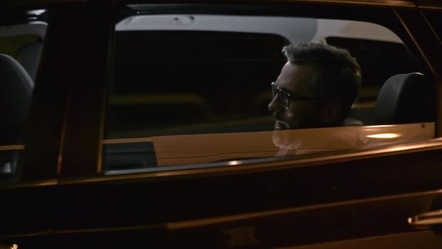 Tracking with side view of successful businessman in glasses and suit drinking coffee while riding in backseat of car driving through city at night