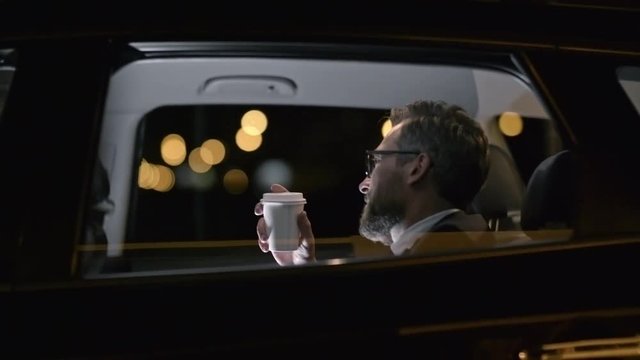 Tracking with side view of bearded businessman in suit and glasses drinking coffee and riding in backseat of car with personal male chauffeur