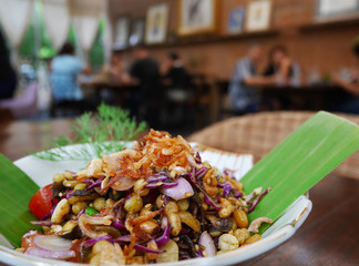 Closeup of Burmese Tea Leaf Salad or Lahpet in a white plate with banana leaf on a dark wooden table with blurry restaurant background