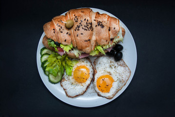 Sandwich Croissant with Fried Eggs. Breakfast with Eggs on White Plate over Black Board. Top view.