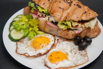 Sandwich Croissant with Cheese, Tomato, Cucumber and Fried Eggs. Breakfast with Eggs on White Plate over Black Board.