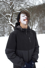 Portrait of a teenager guy in a painted mask, his face is painted black and white. In warm clothes, a hat, a winter day in a snow-covered forest