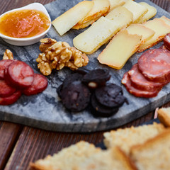 A set of various cheeses, smoked sausage, apricot orange sauce, walnuts, blood sausage and fried bread on a marble board and a wooden table. Square picture.