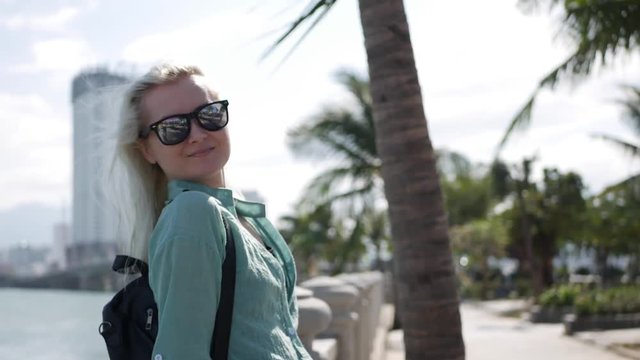 Happy caucasian woman with long blonde hair in sunglasses and green shirt standing and smiling near palm tree on a park and sea background. Travel concept