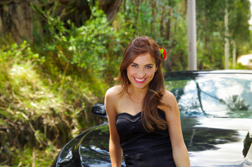 Fototapeta na wymiar Close up of beautiful woman wearing a black dress and a red flower in her head and posing in front of a luxury black car on a road trip, the car standing on the sidelines, in a blurred nature