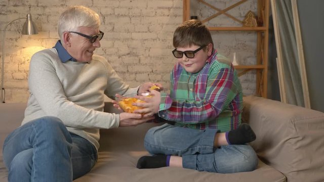 Young greedy boy takes a bowl of chips from his grandfather. Elderly man and a young fat boy are sitting on the couch in 3d glasses. Home comfort, family idyll, cosiness concept, difference of