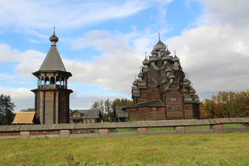 Fototapeta na wymiar The bell tower from logs and traditional Russian church from logs, surrounded with a fencing from logs against the background of the bright autumn sky.