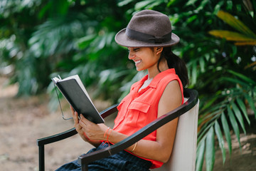 Portrait of a young and attractive Indian Asian girl reading a book in the sun during the day while sitting in a chair in a green park. She is smiling and enjoying herself and is fashionably dressed.	