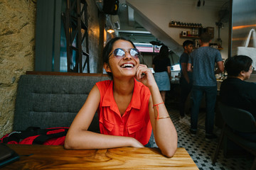 Portrait of an Indian laughing young and fashionable woman sitting in a restaurant during the day. While wearing a pair of sunglasses, she wears a very casual outfit.