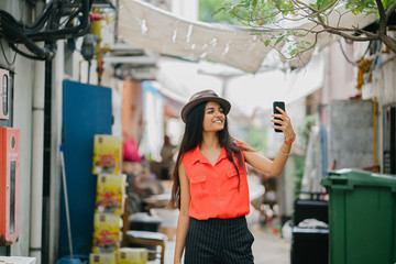 Image of a young and fashionable Indian Asian model wearing a fedora hat while taking a selfie and standing in an alley in Asia. She is smiling and looking classy and is elegant on her outfit.