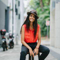 Portrait of an attractive, young and elegant Asian Indian woman sitting in a stool in an Asian alley. She smiles and wears a smart casual with a brown fedora hat.