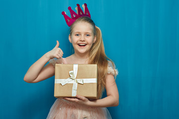 Happy girl wearing pink dress and crown holding gift box showing