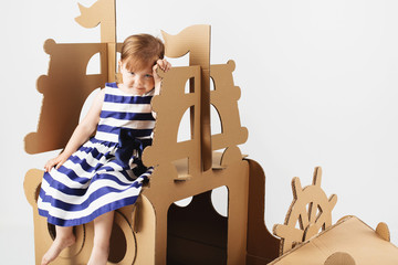 Cute  Little girl wearing striped dress in a marine style playing with cardboard ship on white...