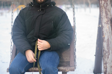 a young man in a jacket and jeans is holding a leash and riding a wooden swing. we return to childhood. adults as children. childhood and rest