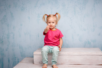 Fototapeta premium Little funny blue-eyed girl child blonde with a haircut two ponytails on her head sitting on a gossip on the background of an old textured wall in blue. Dressed in stylish bright clothes