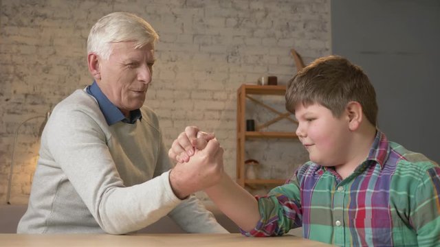 Grandfather and grandson arm wrestle in a cozy room at home. Measuring forces in arm wrestling. Young fat child won. Home comfort, family idyll, cosiness concept, difference of generations, close up