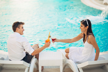 Positive happy couple having a romantic afternoon by the pool in luxury summer vacation resort.Drinking cocktails.Relaxing and enjoying spa wellness weekend.Travel, honeymoon,relationship concept