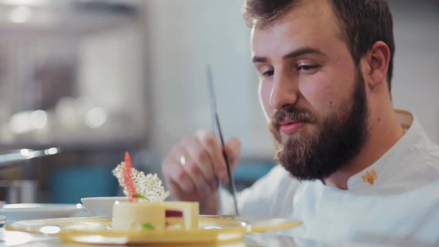 Smiling bearded confectioner decorates the vanilla mousse pastry dessert, checks the result of his work. Professionalism, cheerful mood. Career ladder, motivation. European cuisine, delicious dessert