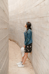 Fototapeta na wymiar Attractive young woman stands between concrete walls. Caucasian girl in casual jeans and flower dress looks up.