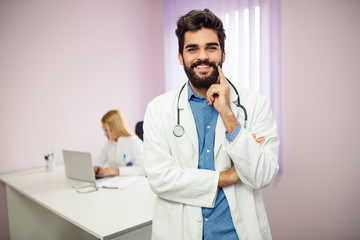 Portrait of a handsome doctor in his office. In the background, his colleague working on a laptop.