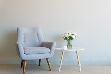 Grey retro armchair with cream roses and cup on small round white table against neutral wall