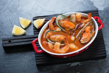 Bowl with buyabes or french fish soup on a black wooden serving board, grey concrete background, horizontal shot