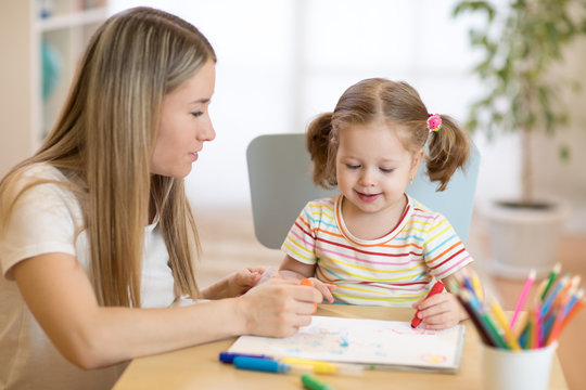 Little child girl coloring with felt pen next to her mother in nursery room.