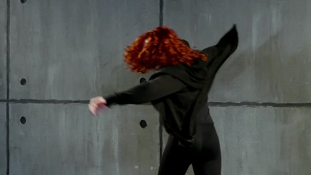 Young red-haired woman performs in black clothes near wall