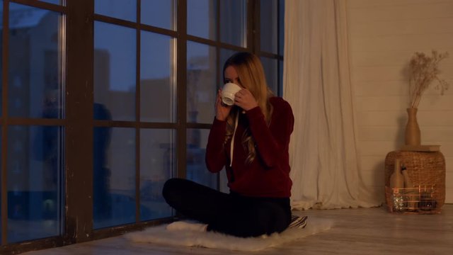 Elegant relaxed young long blonde hair woman enjoying mug of hot coffee and looking out the window dreamily while spending leisure at home in the evening. Hipster girl relaxing in domestic interior.