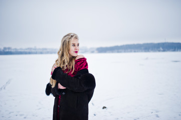 Elegance blonde girl in fur coat and red evening dress posed at winter snowy day.