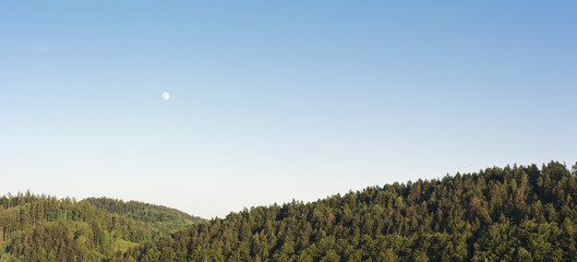 Fototapeta premium View of forested hills with moon in the sky during the day.