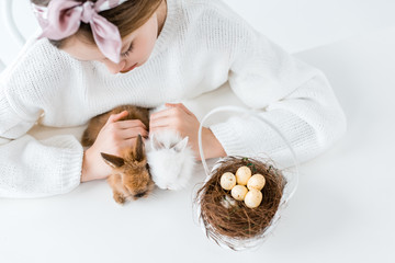 Fototapeta na wymiar cropped shot of girl looking at cute furry rabbits and basket with easter eggs in nest