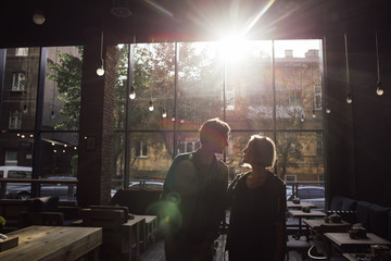 Fototapeta na wymiar Nice couple standing on cozy empty cafe in loft style looking to each other with sunlight back view