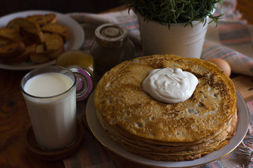 A stack of delicious fresh thin pancakes with sour cream on a table covered with a tablecloth and a glass of milk cooked for Maslennitsa's day.