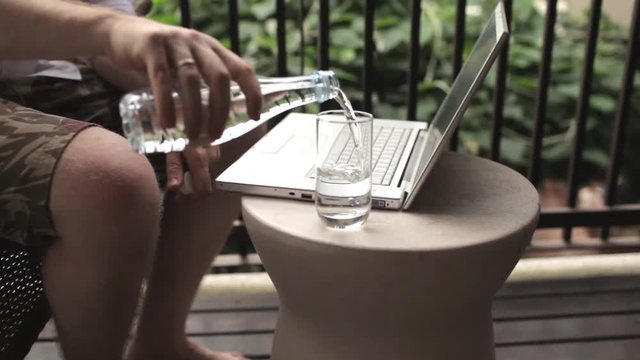 man with a laptop pours clean water in a glass sitting on the terrace